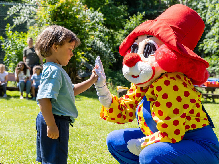 Kind interagiert mit Familotel Maskottchen Happy auf einer grünen Wiese