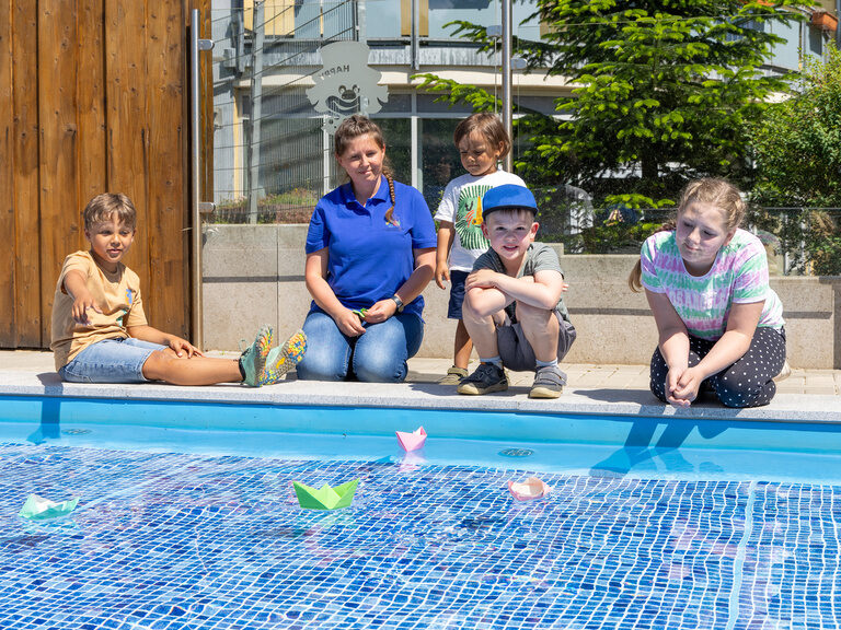 Kinder und Betreuer sitzen am Rand des Pools und spielen mit Wasserspielzeug